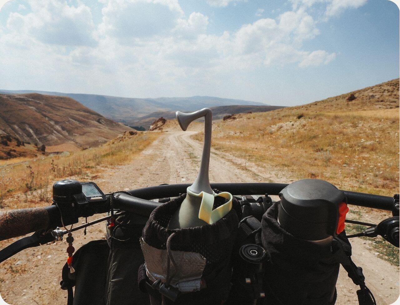 Bidetlity Po-Dusche in Gentle Grey sicher verstaut auf einem Fahrradlenker, bereit für ein Abenteuer auf einer staubigen Offroad-Strecke durch eine weite Hügellandschaft. Perfekt für hygienische Frische unterwegs